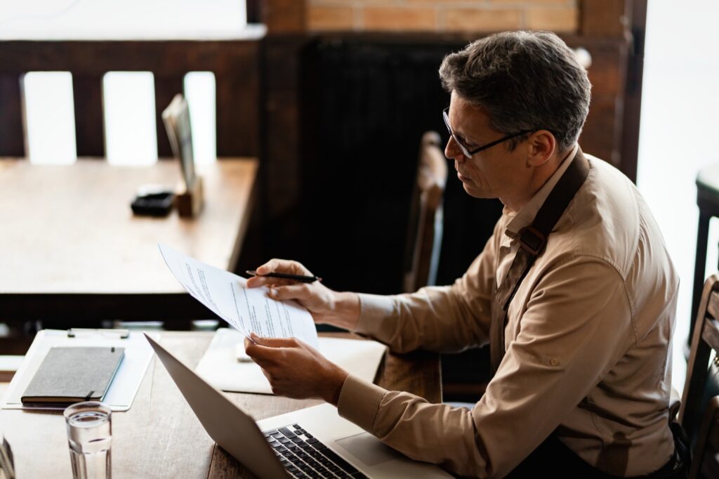 Dono de um pequeno negócio analisando relatórios impressos ao lado de um notebook, sentado a uma mesa de café